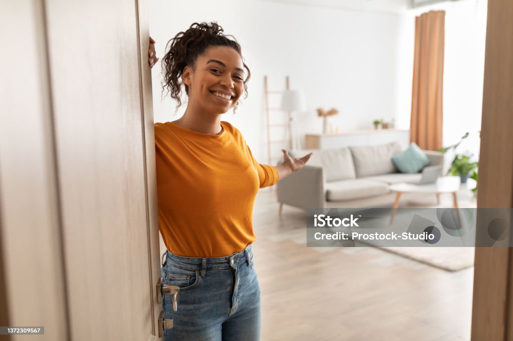 Cheerful African Woman Opening Door Welcoming You Standing At Home Cheerful African American Woman Opening Door And Gesturing Welcoming You To Come In Smiling To Camera Standing At Home. Hospitality, Real Estate Ownership And Purchase Concept Home Ownership Stock Photo