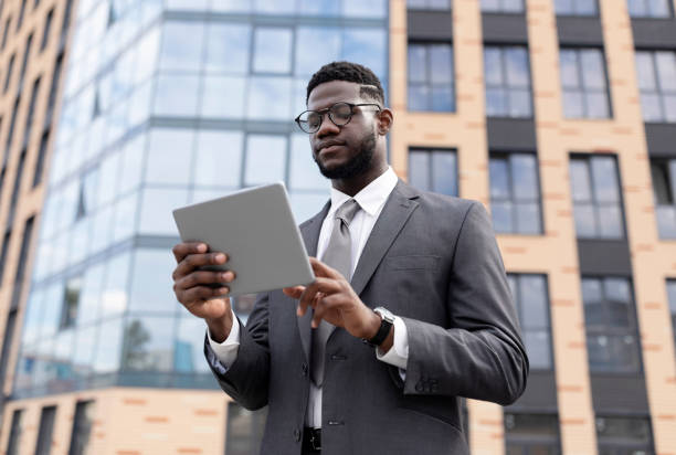 istock Focused black man in formal wear standing by office building, holding digital tablet, using business application 1372309927