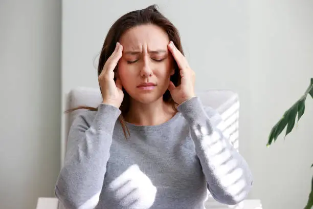 Photo of Shot of a young woman holding her head in discomfort due to pain at home