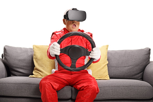 Matrue man in a car racing suit wearing vr headset and holding a steering wheel seated on a sofa isolated on white background