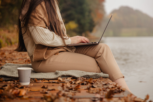 Young woman working on a laptop on the wooden platform by the lake on a cloudy day in autumn.