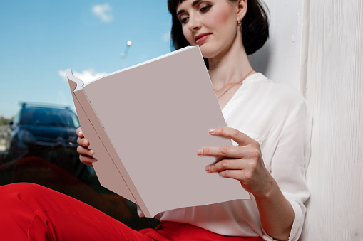beautiful young woman holding a magazine with mock up. girl in a white shirt and red pants sitting near the window.