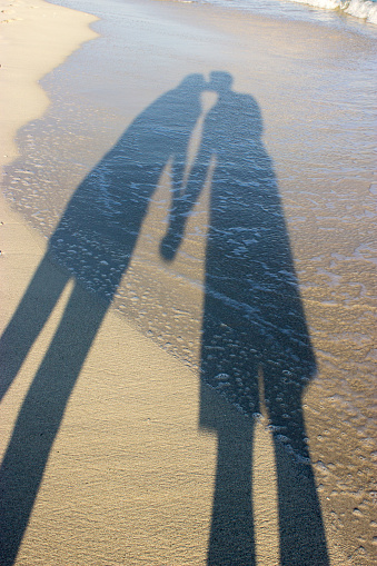 Shadows of a loving couple kissing on the sand of the beach bathed by the sea at dawn, lifestyle concept. Vertical