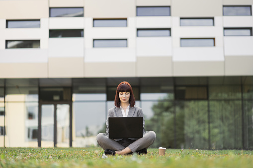 Distance learning, online education and work outside. Confident attractive young woman in formal wear, sitting on green grass with legs crossed and using laptop outside office, during coffee break