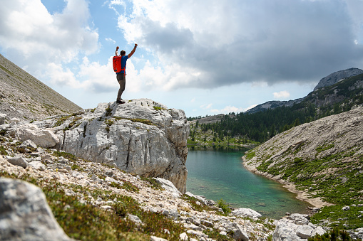 Young man standing on a big rock overlooking beautiful mountain lake. Handing hiking poles and with hands in the air.