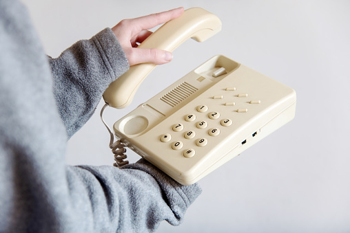 Detail of a woman's hand picking up an old white corded telephone. Hand holding the receiver of a home or office landline phone. Concept of answering a phone call.