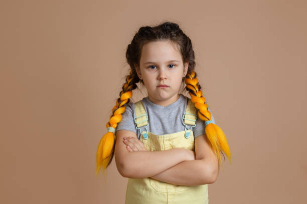 黄色いカネカロンの三つ編みを持つ若い気分を害した白人の子供は、黄色のジャンプスーツとベージュの背景に灰色のtシャツで腕を組んで、不快感を示す - anger child braids braided ストックフォトと画像