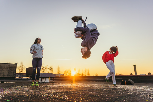 Young man perform backflip with women dancing in background on roof during sunset