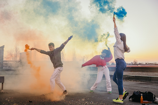 Young friends dancing and holding multi colored smoke bombs on roof during sunset