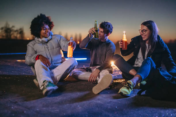 Young friends with drinks having good time while sitting on rooftop during dusk
