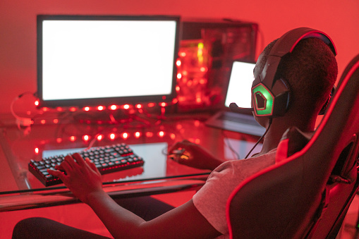 A young black male gamer is playing at his station using desktop computer and laptop