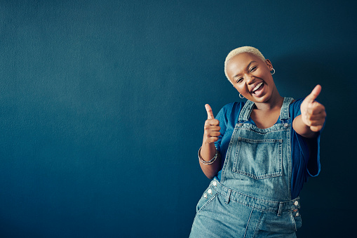 Portrait of a laughing young woman in a blue t-shirt and overalls giving two thumbs up in front of a blue background