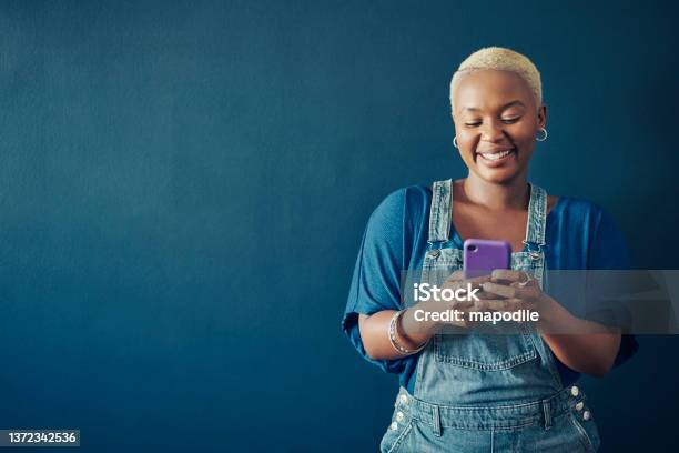 Smiling Woman In Overalls Texting On Her Phone Against A Blue Background Stock Photo - Download Image Now