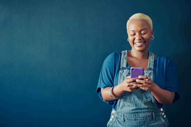 smiling woman in overalls texting on her phone against a blue background - women smiling blond hair human face imagens e fotografias de stock
