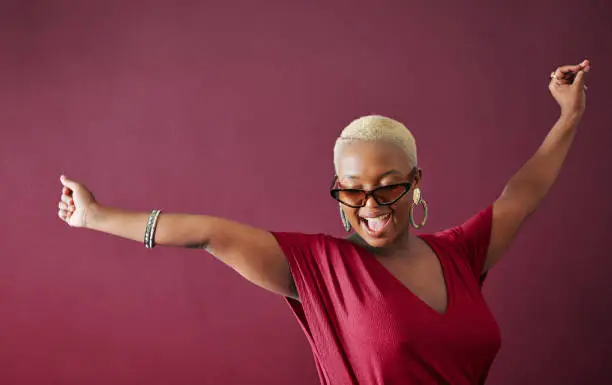 Carefree young woman in a red blouse and sunglasses celebrating with her arms raised and laughing in front of a red background