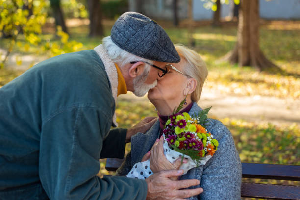 älterer mann, der seine frau im park küsst. ehemann schenkt seiner frau blumen - senior adult women park bench 70s stock-fotos und bilder