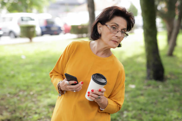 Portrait of a old woman 70-75 years old, with a phone and a cup for single use with coffee and is walking in a city park stock photo