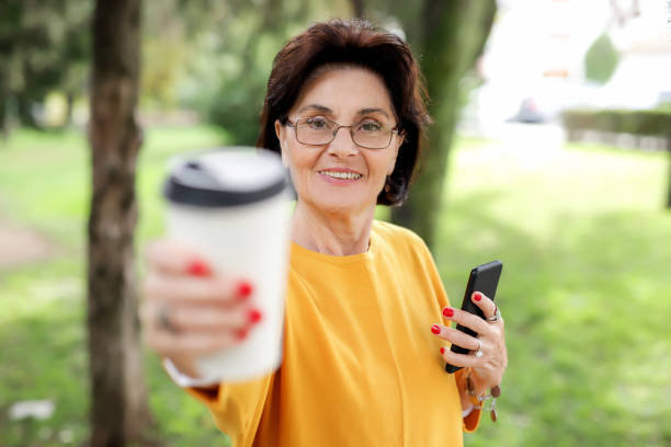 Portrait of a smiling old woman 70-75 years old, with a phone and a cup for single use with coffee and is happy walking in a city park stock photo