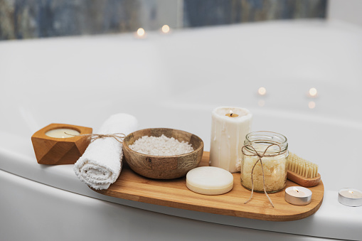 Preparation for hotel spa treatment, home bath procedure. White washbasin in bathroom, accessories on tray. Burning candles, soap, foot brush, towel, glass bottle with sea salt, orchid flower