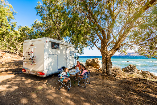 Family traveling with motorhome are eating breakfast on a beach. Travelers on an active family vacation with motorhome RV parked on the beach under a tree facing the sea, Crete, Greece.