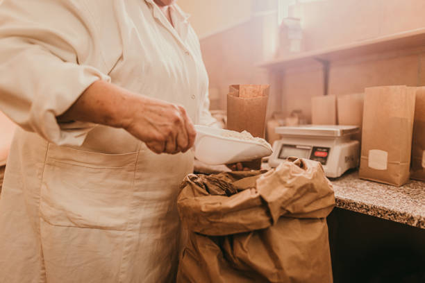 Senior woman packing fresh flour in paper bag with scoop Person in uniform taking fresh flour with scoop for packing in brown paper bag beside table flour mill stock pictures, royalty-free photos & images
