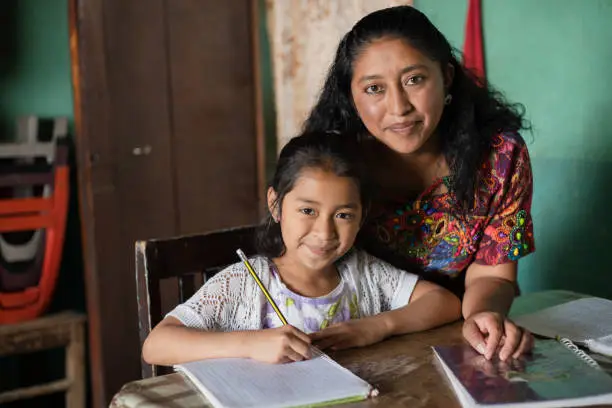 Photo of Hispanic mom helping her little daughter do her homework - Mom teaching her daughter to read and write at home - Mayan family at home
