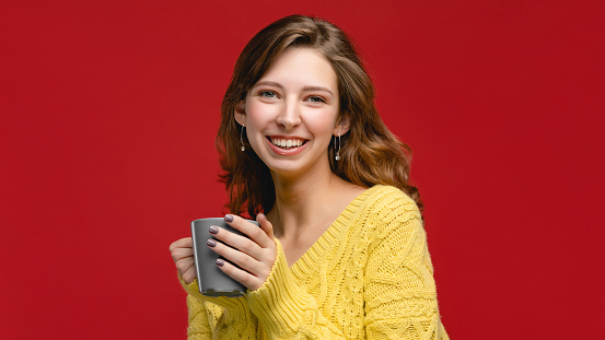Beautiful woman in studio is holding and drinking beverage from mug. Concept with people on red background, person wear yellow jumper and smiling. Girl with coffee or tea in the cup