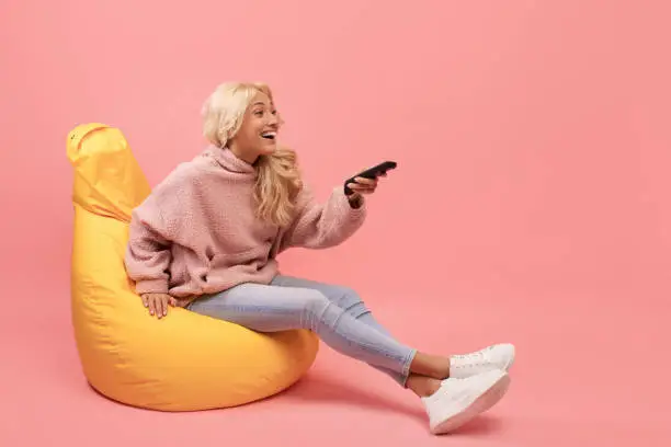 Excited lady with remote control sitting in beanbag chair and watching TV against pink background, free space. Young woman with television controller switching television channels