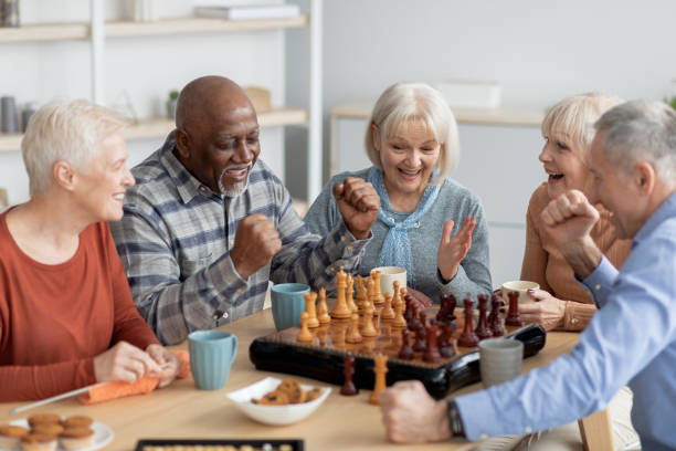 Emotional multiethnic group of senior people playing chess Emotional multiethnic group of senior people playing chess, happy elderly men and women in casual outfits sitting around table with table games, chatting, having fun at sanatorium senior chess stock pictures, royalty-free photos & images