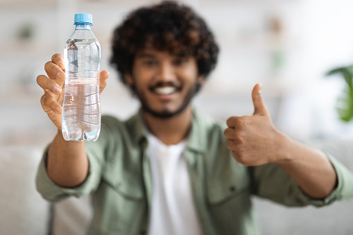 Happy curly young indian man drinking fresh water and smiling, guy showing bottle of water and thumb up, body hydration concept, selective focus on bottle, closeup photo, blurred background