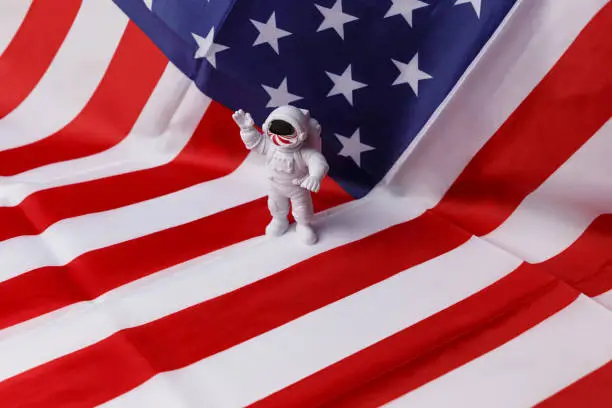 The concept of the Cosmonautics Day. A toy astronaut waves his hand and stands against the background of the national flag of America. Space patriot. Side view.