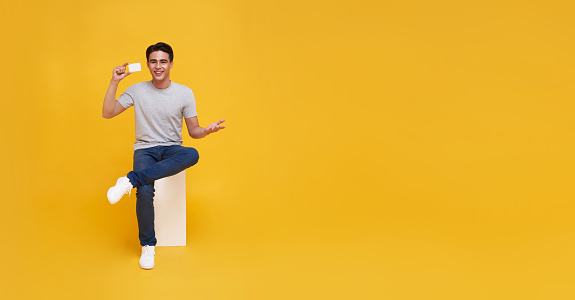 Happy handsome Asian man showing credit card for making payment or paying online business in hand isolated on yellow background.