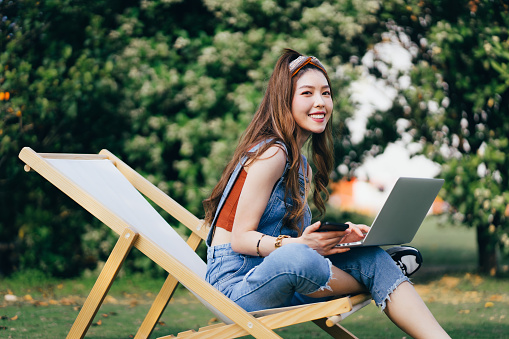 Portrait of beautiful stylish young Asian woman holding smartphone, using laptop while sitting on deck chair on a meadow outdoors in park. Cheerful young female in denim looking at camera. Enjoying the sun and fresh air while using laptop outdoors