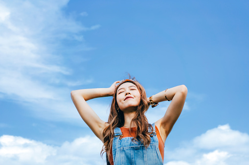 Beautiful young Asian woman with eyes closed open arms and taking deep breath outdoors in nature, against blue sky on a sunny day. Enjoying sunshine and freedom in nature