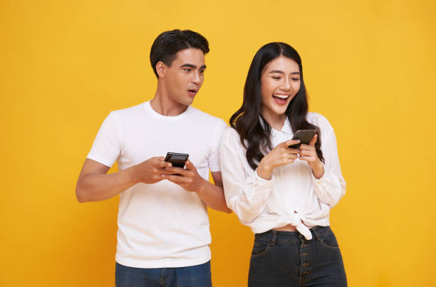 istock Shocked asian man spying on her smiling girlfriend while both using mobile phones isolated over yellow background. chatting or social media concept. 1372360627
