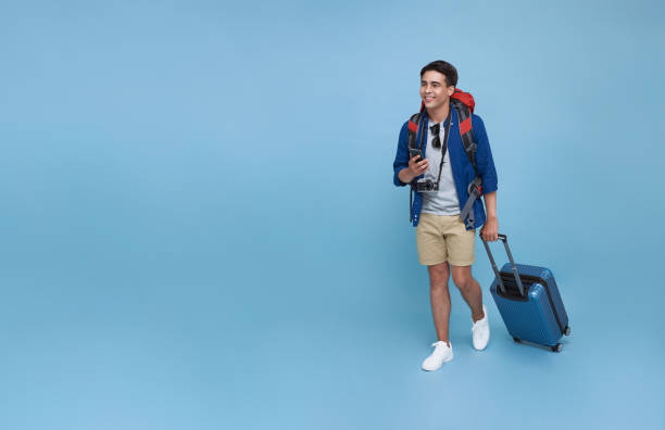 Happy young Asian tourist man holding smartphone with baggage going to travel on holidays isolated on blue copy space background. stock photo