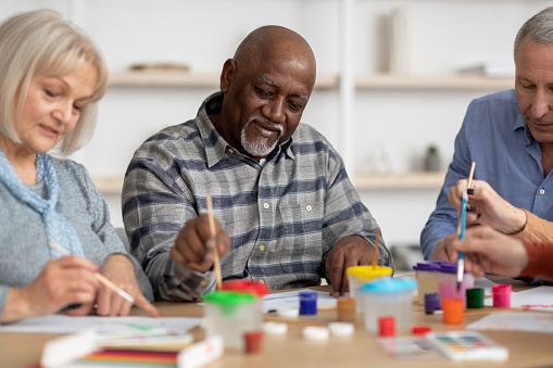 Art therapy for seniors concept. Happy multiracial elderly men and women enjoying painting with brush, having conversation and smiling, attending drawing master-class together