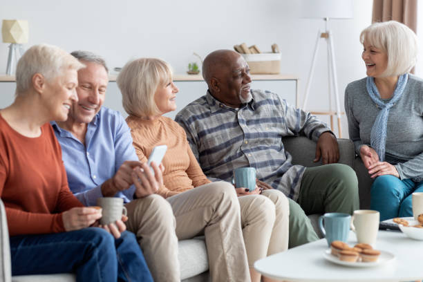 International group of cheerful elderly people chilling together International group of cheerful elderly people men and women in casual outfits chilling together at sanatorium, sitting on sofa in living room, using mobile phone, drinking tea, chatting aging population stock pictures, royalty-free photos & images