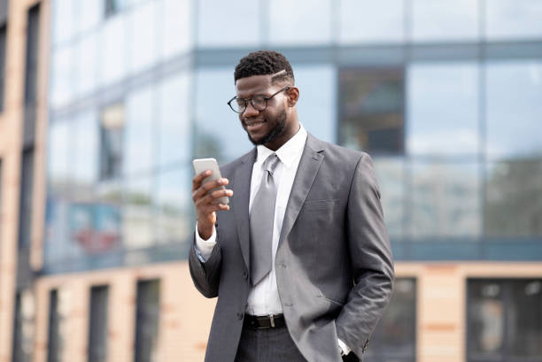 istock Successful black IT manager in formal suit walking next to office building, using smartphone and smiling, copy space 1372360943