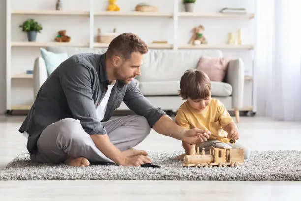 Photo of Happy young father spending time with his cute little son at home, sitting on floor and playing wooden train, free space