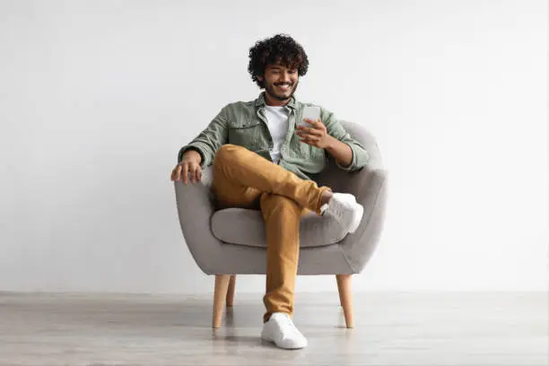 Happy handsome curly young indian man chatting with girlfriend while relaxing in armchair at home, using modern mobile phone, checking social media, using mobile app, panorama with copy space