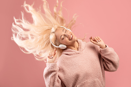 Enjoy the music concept. Joyful lady listening to music in wireless headphones and dancing, playing with hair over pink background. Young woman enjoying playlist