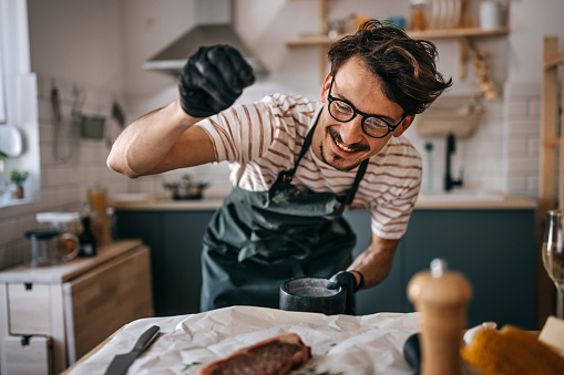 Happy young man wearing eyeglasses sprinkling pepper on steak in kitchen at home