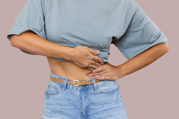 Young woman holding her side with her hands. Sore side under the rib after training. Stomach pain Cropped shot of a young woman in a grey t-shirt and denim shorts holding her side with her hands isolated on color beige background. Sore side under the rib after training. Stomach pain gall bladder stock pictures, royalty-free photos & images