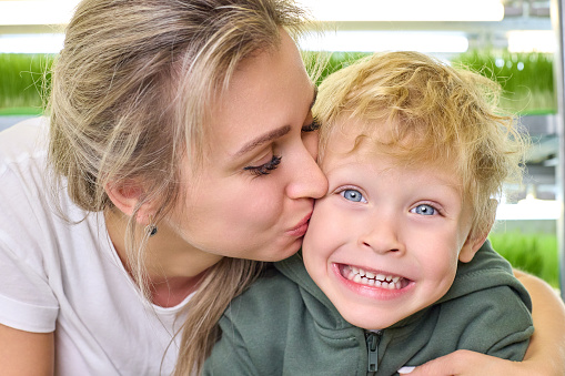 Young beautiful mother kisses her happy son against the background of bright shelves with micro greenery. Concept of the influence of proper nutrition on family health and human happiness