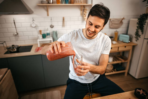 istock Smiling man pouring smoothie in glass while sitting in kitchen 1372373577
