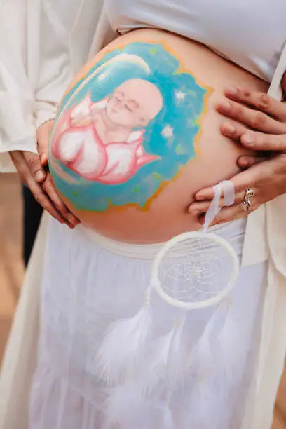 Body part of a pregnant woman with artistically painted belly with a baby mandala and the future father embracing her belly. Selective focus, color editing and added grain. Real life situation. Part of a series.