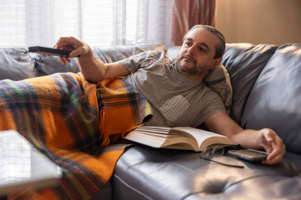 istock Feeling bad. A mature man is ill, rest on a couch under a warm plaid. He is use a remote to switch TV chanels. 1372380981
