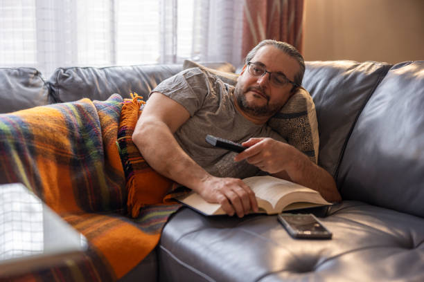 istock Feeling bad. A mature man is ill, rest on a couch under a warm plaid. He is use a remote to switch TV chanels. 1372380989