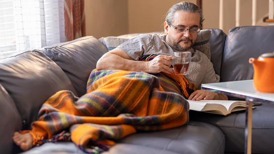 A sick mature man is resting on a couch covered by a plaid, resting during his ilness.